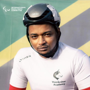 Hilmy Shawwal wearing a helmet and a white athletic shirt that reads Weir Archer Academy. In the background the Tanzanian flag and in the top left corner the Paralympic Games logo.