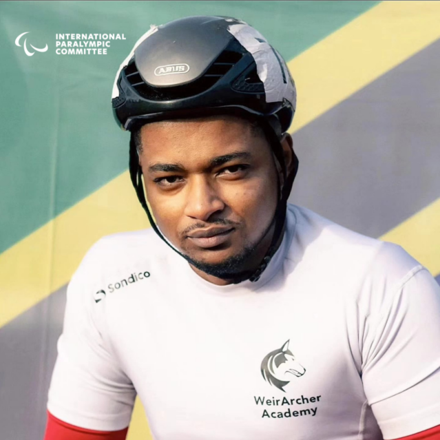 Hilmy Shawwal wearing a helmet and a white athletic shirt that reads Weir Archer Academy. In the background the Tanzanian flag and in the top left corner the Paralympic Games logo.