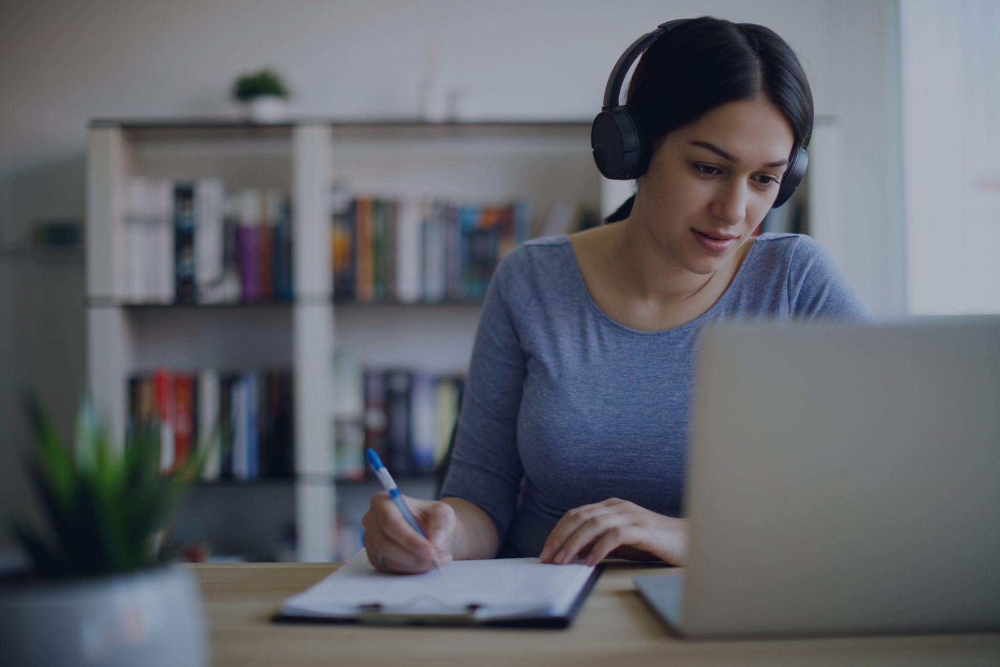 A woman waring headphones looking at a laptop while writing notes on a clipboard.