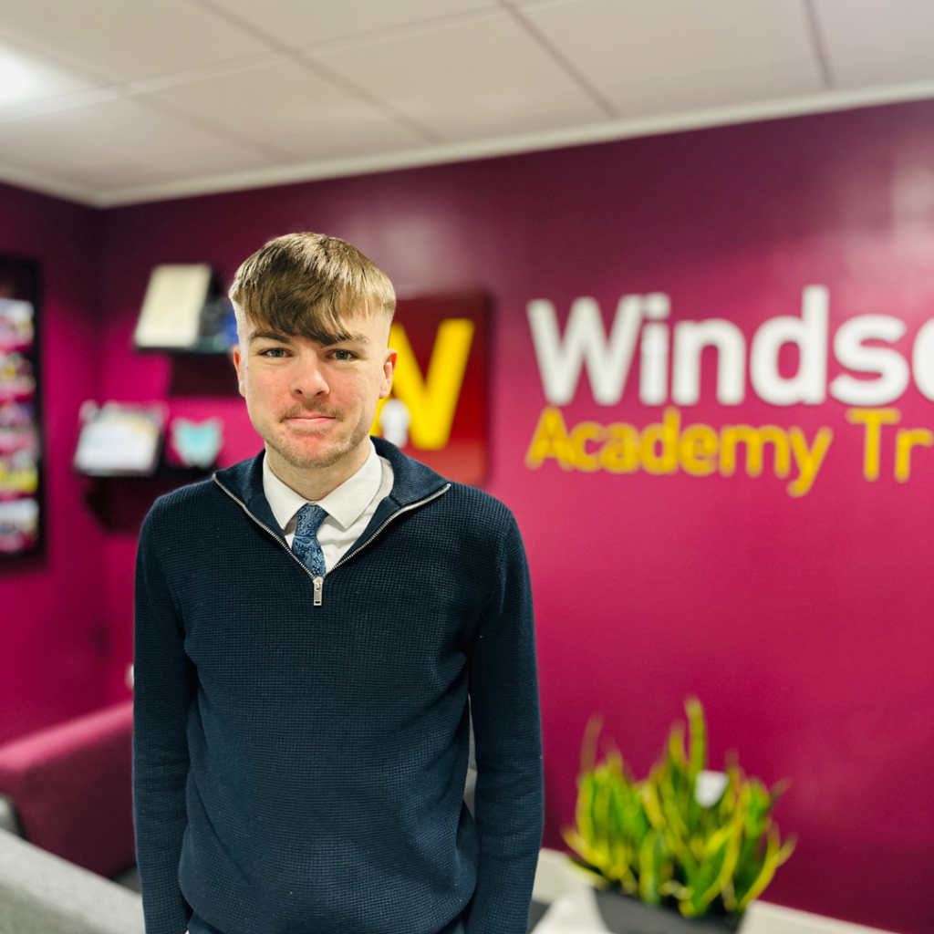 Photo of Joel looking at the camera. Joel is wearing a jumper and a shirt and tie underneath. In the background a red wall with the Windsor Academy Trust name and logo.