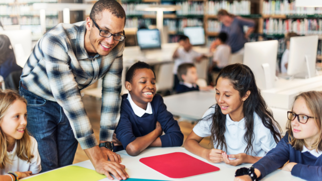 Teacher standing over a table with 4 pupils. The teacher's hand is on the table showing that he is offering help.