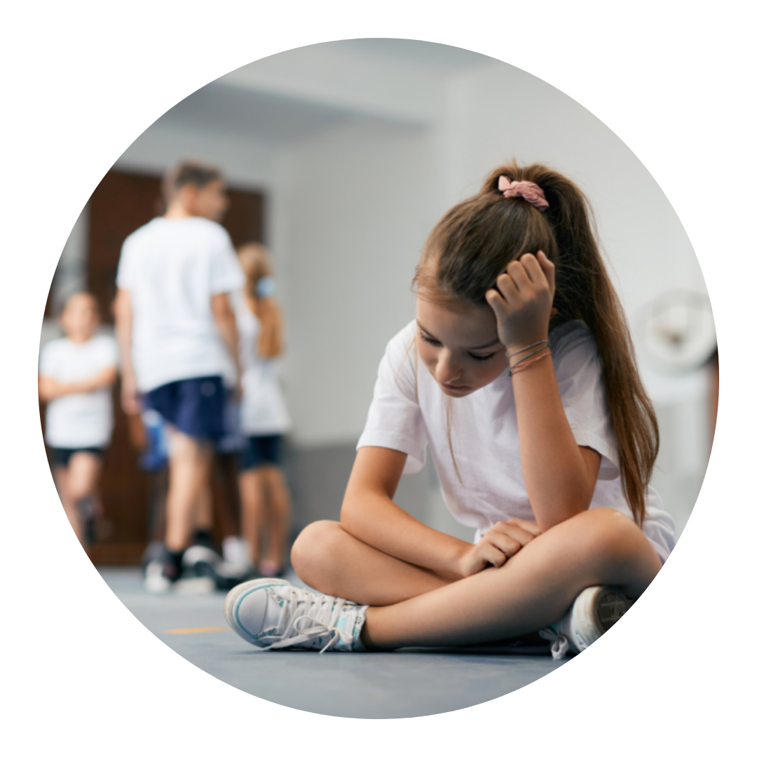 Young girl sitting on the floor crossed legged with her head leaning on her hand. In the background other pupils who are not engaging with her.