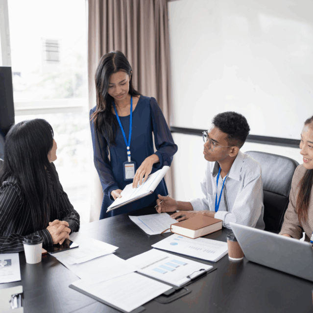 Three people around an office desk, one woman standing and the other three people looking at her attentively as she points at a clikboard. Other clikboards with pages showing various graphs scattered on the desk in front of them.