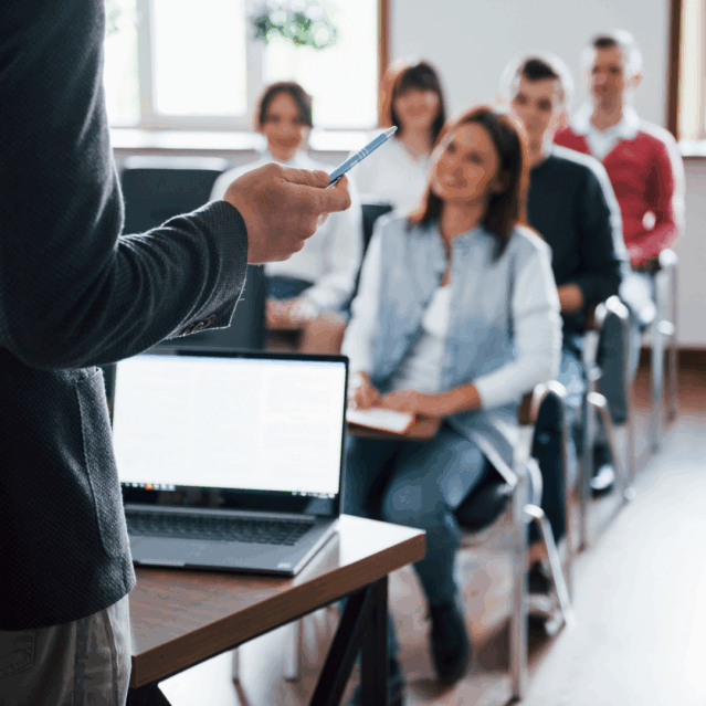 In the foreground a back of a person teaching a class, an open laptop in front of them. In the background several students looking at the teacher in concentration.