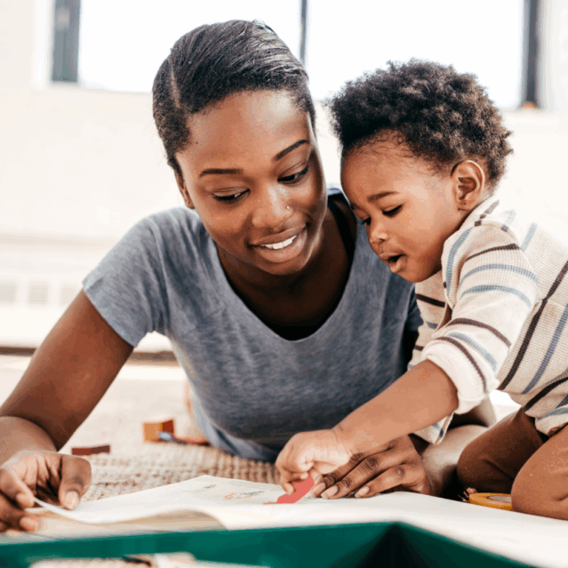 A woman smiling as she holds a toddler and shows him a book.
