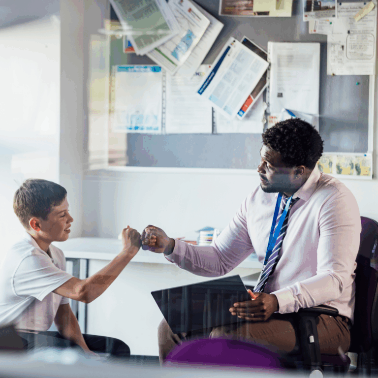 A young teacher gives a fist bump to a smiling pupil in school uniform.