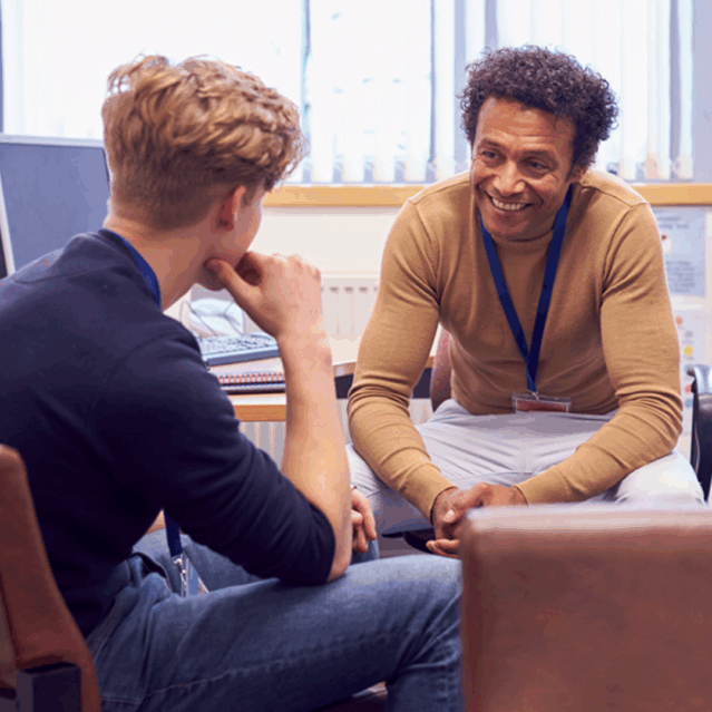 A pupil and teacher in an office speaking to each other. The pupil has his back to us and appears to be listening to the teacher, the teacher is smiling at the pupil.