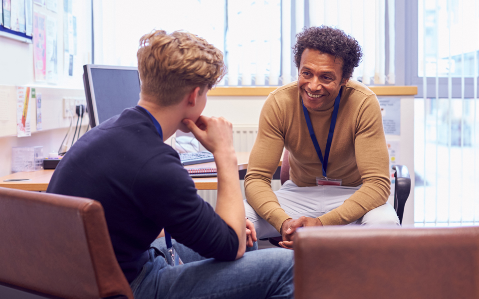 A pupil and teacher in an office speaking to each other. The pupil has his back to us and appears to be listening to the teacher, the teacher is smiling at the pupil.