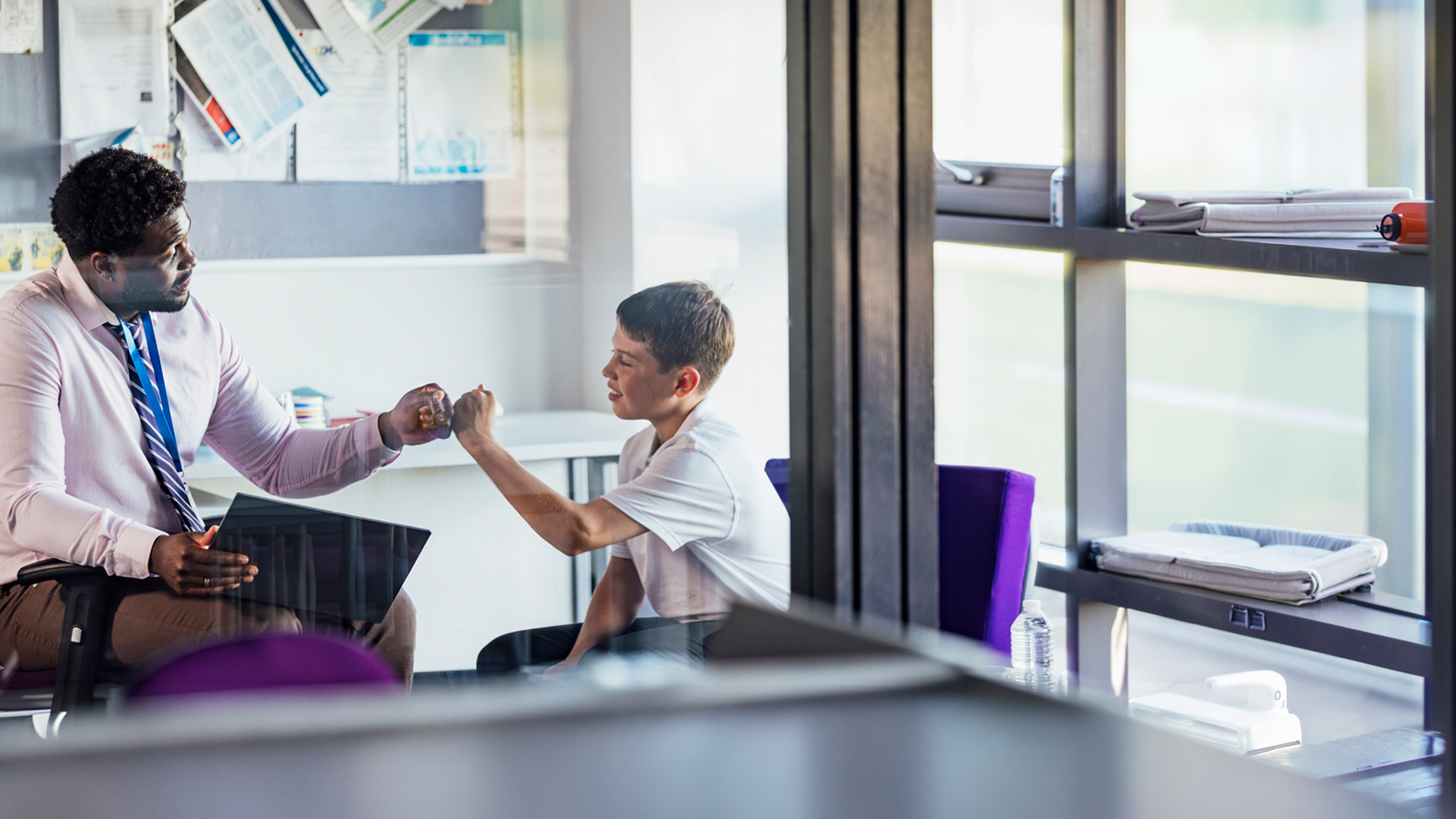 A young teacher gives a fist bump to a smiling pupil in school uniform.