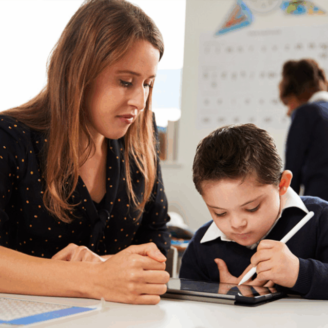 A young pupil concentrating as he works with an iPad. Next to him a female teacher looking at his work attentively.